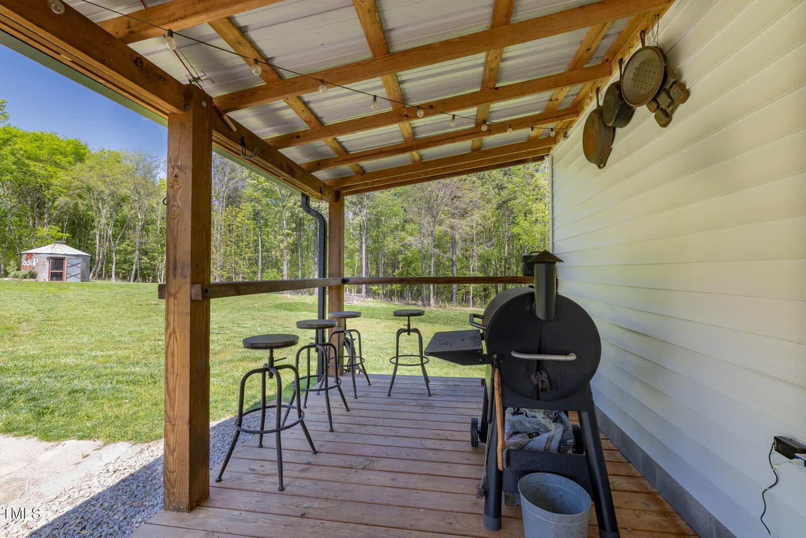 A covered wooden deck with a metal grill, three bar stools, and a small counter overlooking a grassy yard. Metal pots and pans hang on the house's white siding. Trees and a shed are visible in the background.