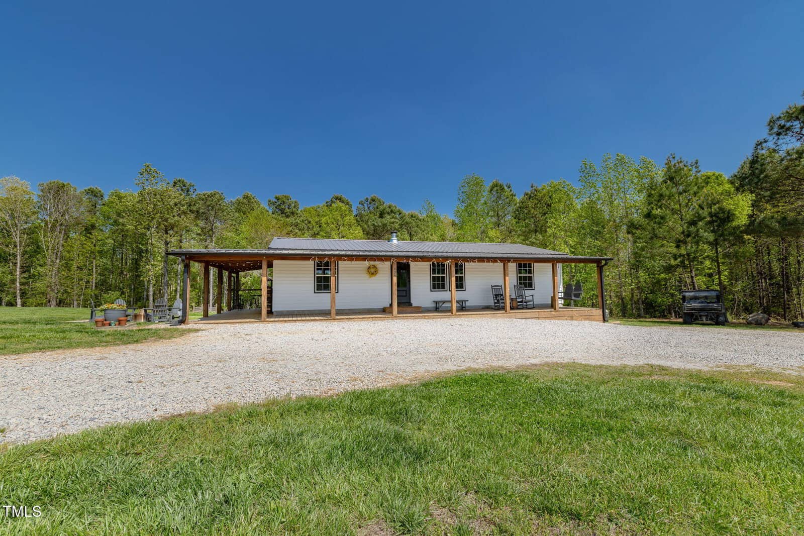 A single-story white house with a covered front porch sits in a rural area, surrounded by green grass, trees, and a gravel driveway under a clear blue sky. Outdoor seating and vehicles are visible near the house.