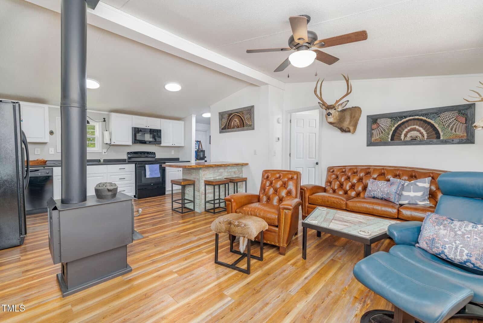 A living room with wood floors features brown leather chairs, a blue recliner, a wood stove, and a glass coffee table. The kitchen with white cabinets and a black fridge is visible. Deer mounts and turkey fans are displayed on the walls.