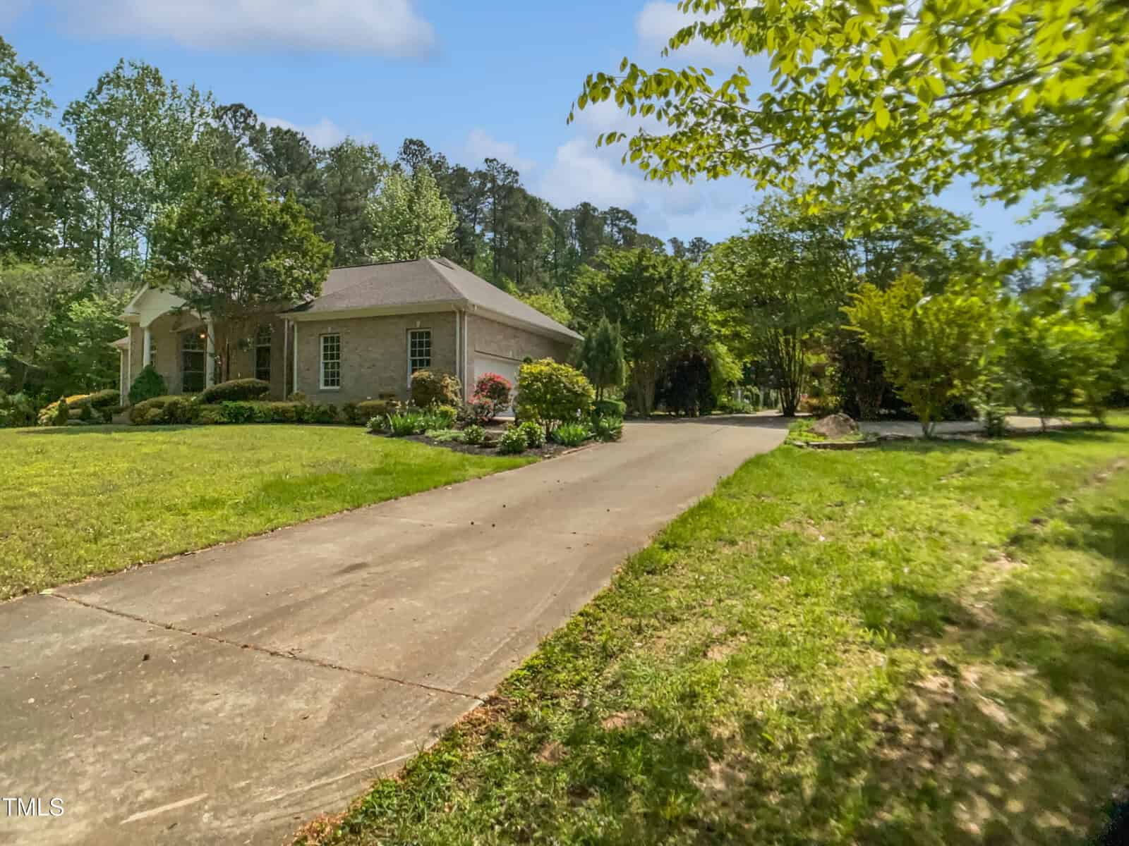 A light-colored single-story house with a front porch sits on a grassy lawn, surrounded by trees and shrubs, with a long concrete driveway leading up to it under a partly cloudy sky.