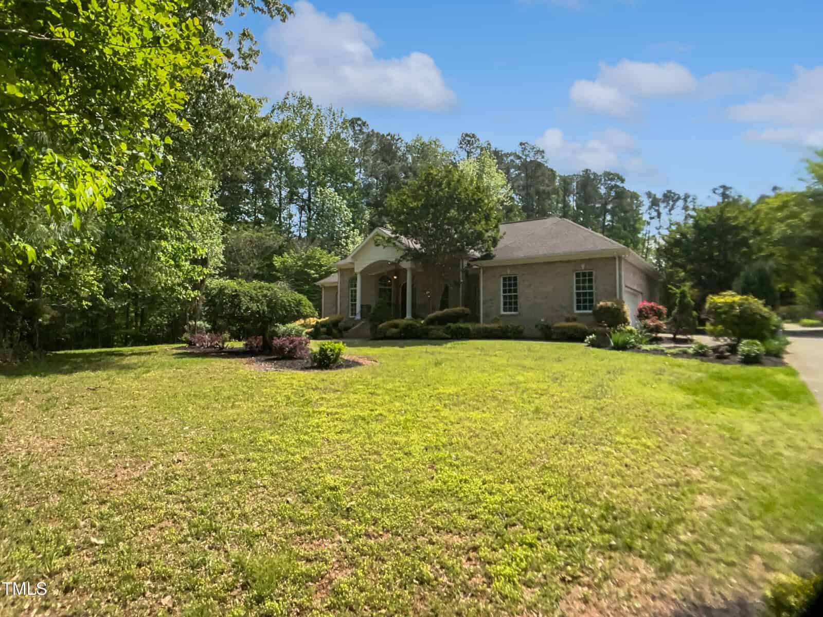 A single-story house with a brick exterior sits surrounded by trees and landscaped bushes, with a well-kept lawn under a blue sky with scattered clouds.