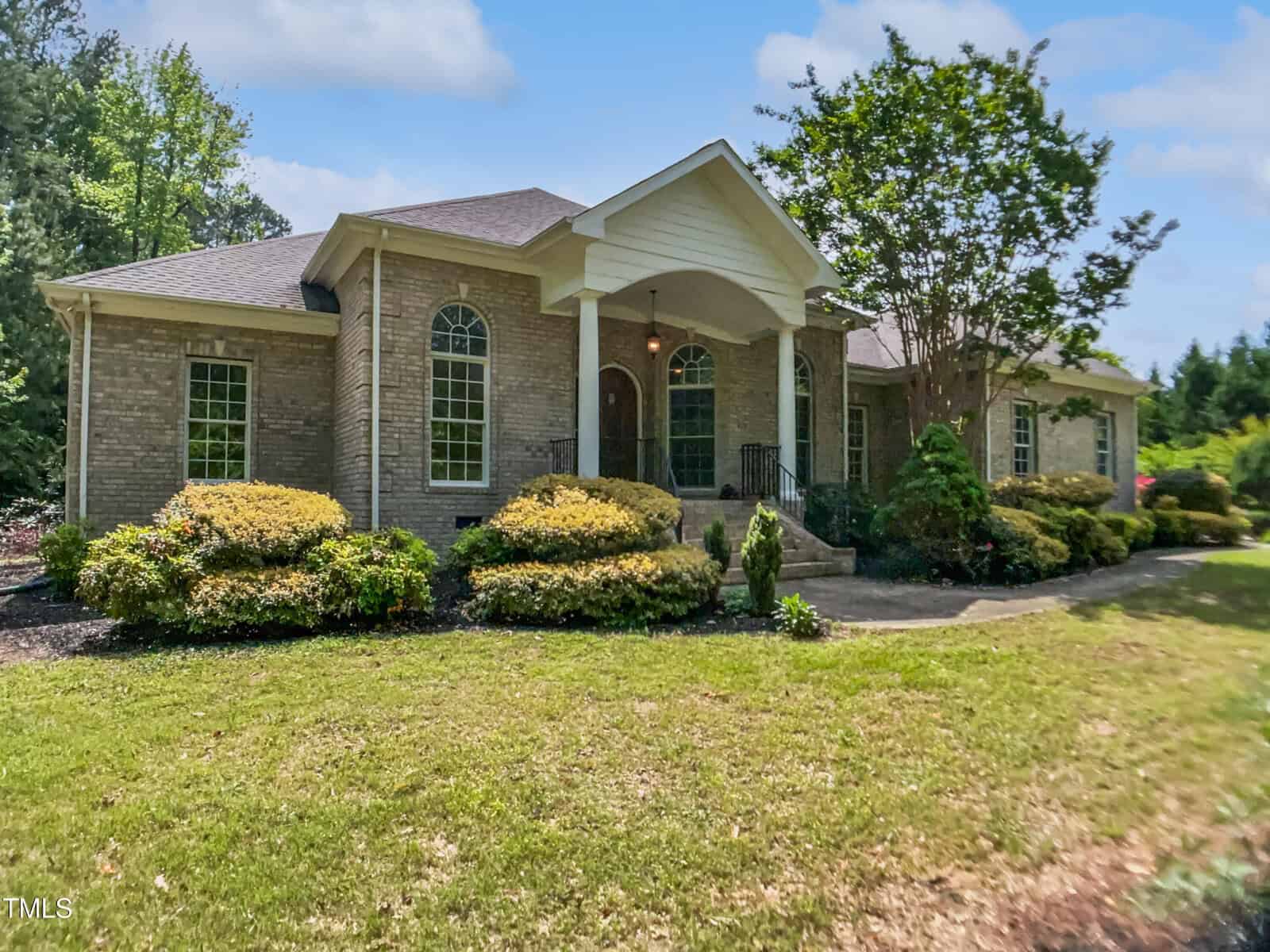 Single-story brick house with arched windows, covered front porch supported by columns, landscaped shrubs, and a neatly maintained lawn on a sunny day.