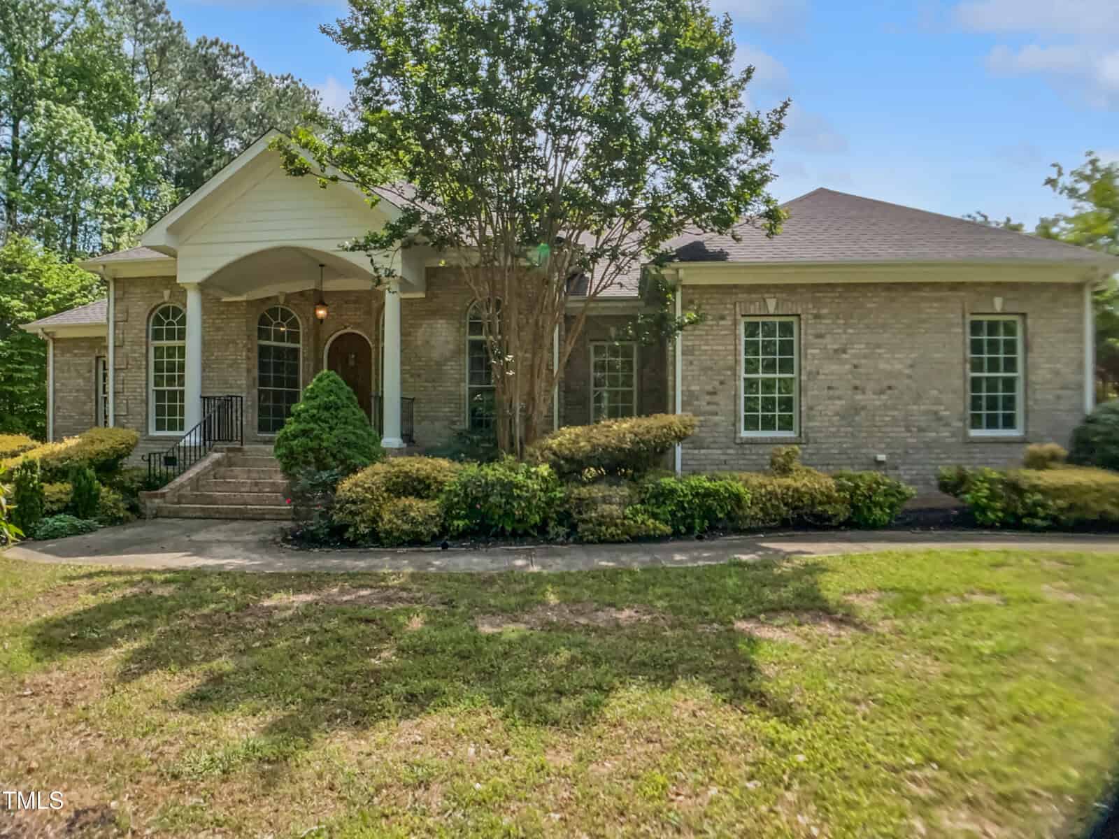 A single-story brick house with large arched windows, white columns by the entrance, trimmed bushes, and tall trees in the front yard on a sunny day.