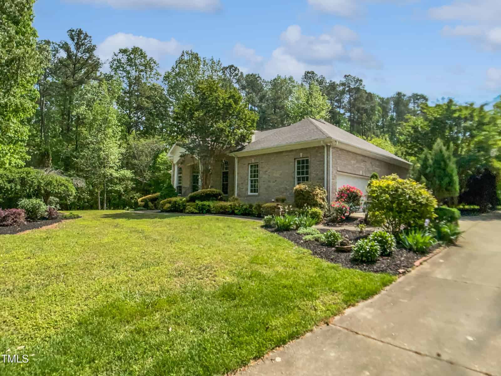 Single-story brick house surrounded by lush landscaping, shrubs, and trees, with a well-maintained lawn and a driveway on the right side, set against a backdrop of tall trees under a blue sky with clouds.