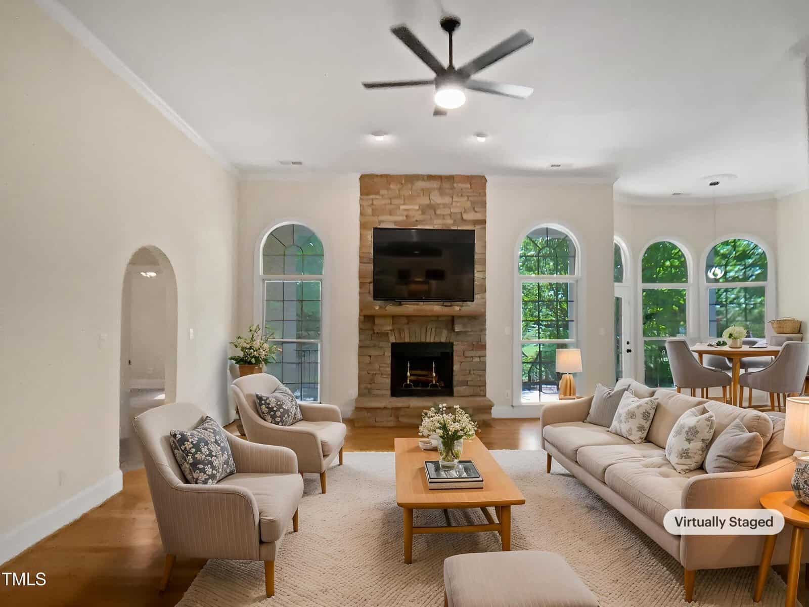 A bright living room with beige walls, a stone fireplace, wall-mounted TV, light-colored sofas and chairs, wood flooring, large windows, and a dining nook in the background. The image is virtually staged.