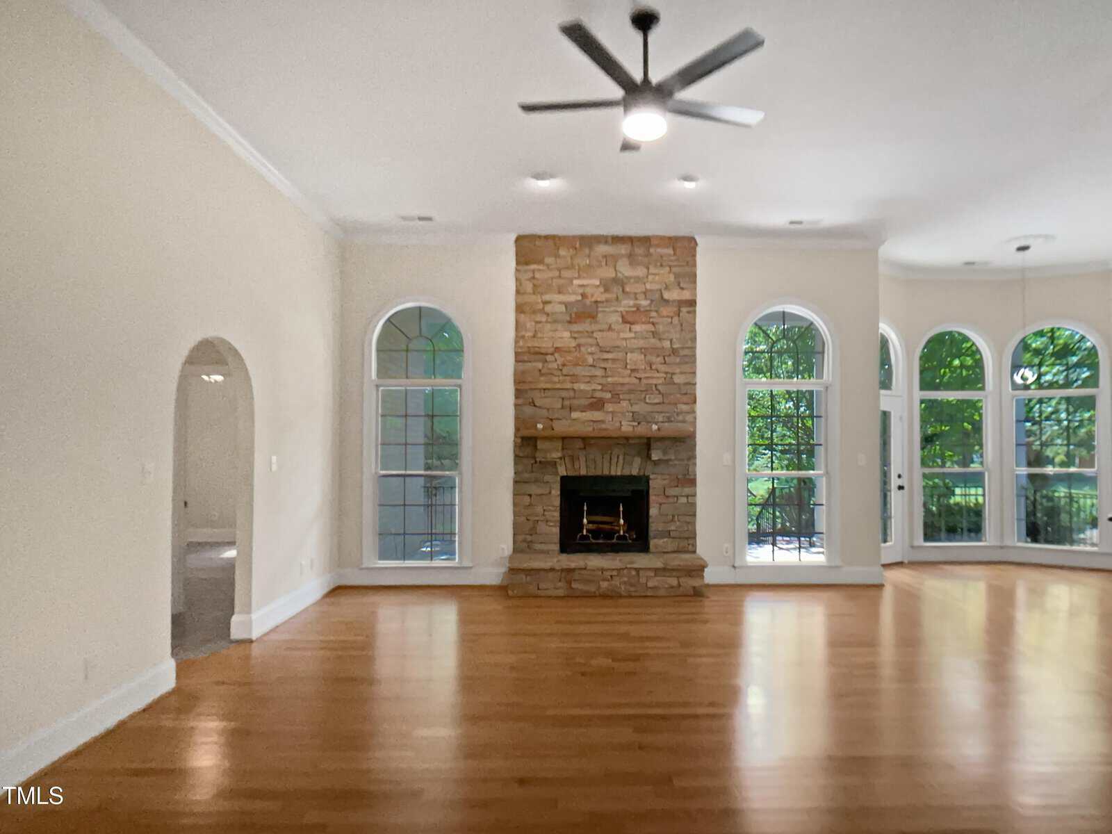 Spacious living room with light wood floors, a stone fireplace centered on the back wall, tall arched windows on either side, and a ceiling fan with light. Natural light fills the room from the windows.