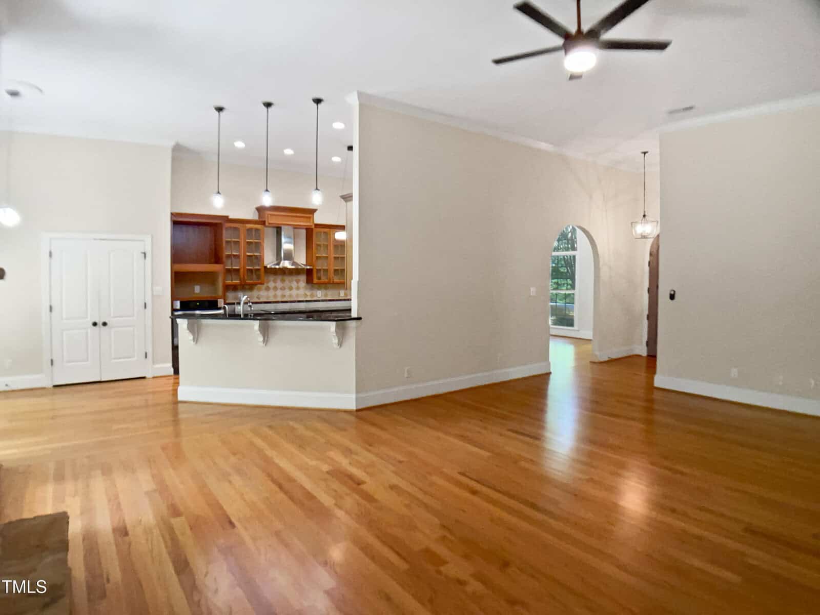 Open living space with hardwood floors, beige walls, ceiling fan, and recessed lights. The kitchen has wooden cabinets, a tile backsplash, and a breakfast bar. An arched doorway leads to another room with large windows.
