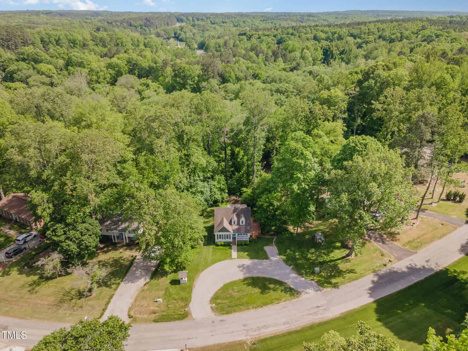 Aerial view of a two-story house with a circular driveway, surrounded by dense green trees and neighboring homes, located on a curved road in a forested area.
