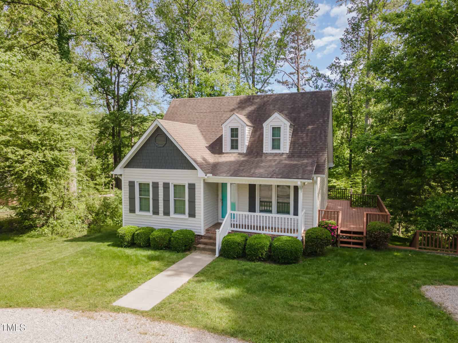 A white, single-story house with dormer windows, a covered front porch, and a wooden deck on the right side, surrounded by green lawn, bushes, and trees under a partly cloudy sky.