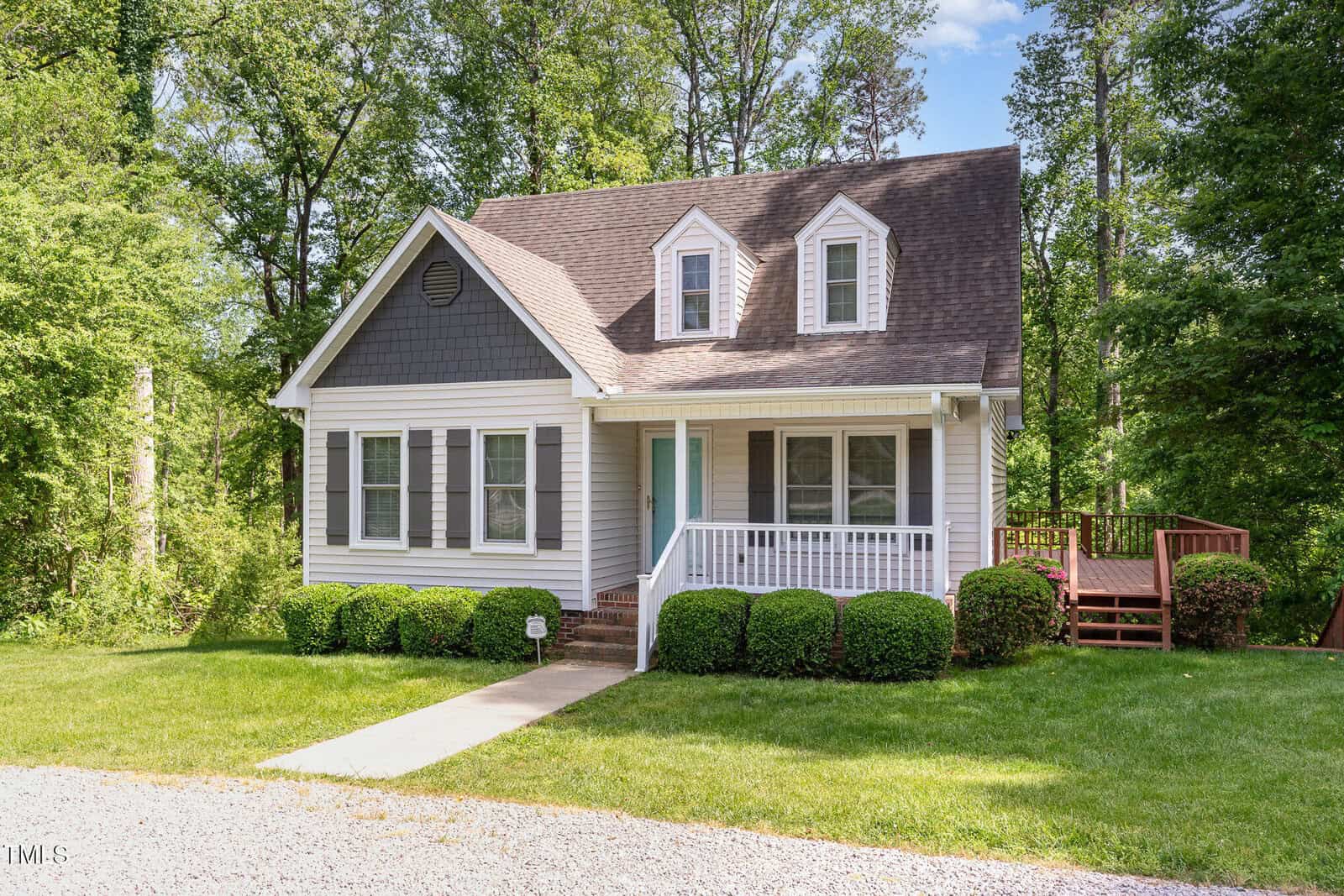 A small white house with a dark gray gable, three dormer windows, front porch, and a side wooden deck. The house is surrounded by green shrubs, trees, and a lawn, with a gravel driveway in front.