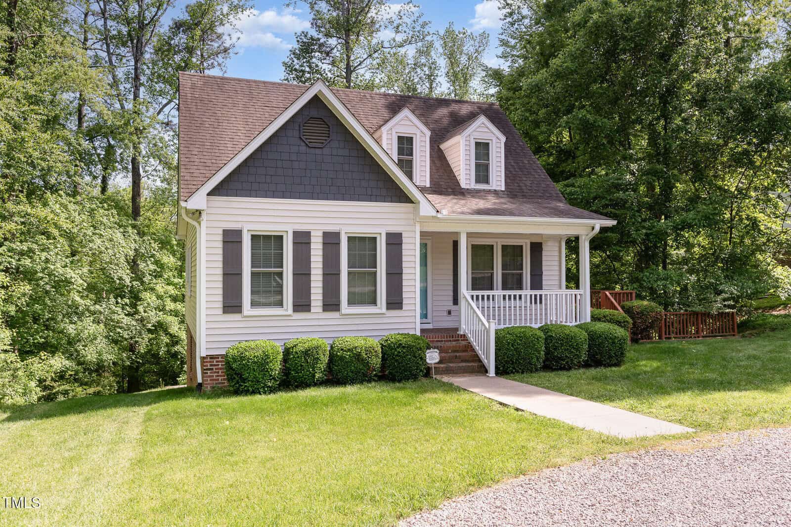 A small two-story house with blue-gray siding, white trim, and a covered front porch sits on a well-maintained lawn. Shrubs line the front of the house, and trees are visible in the background.