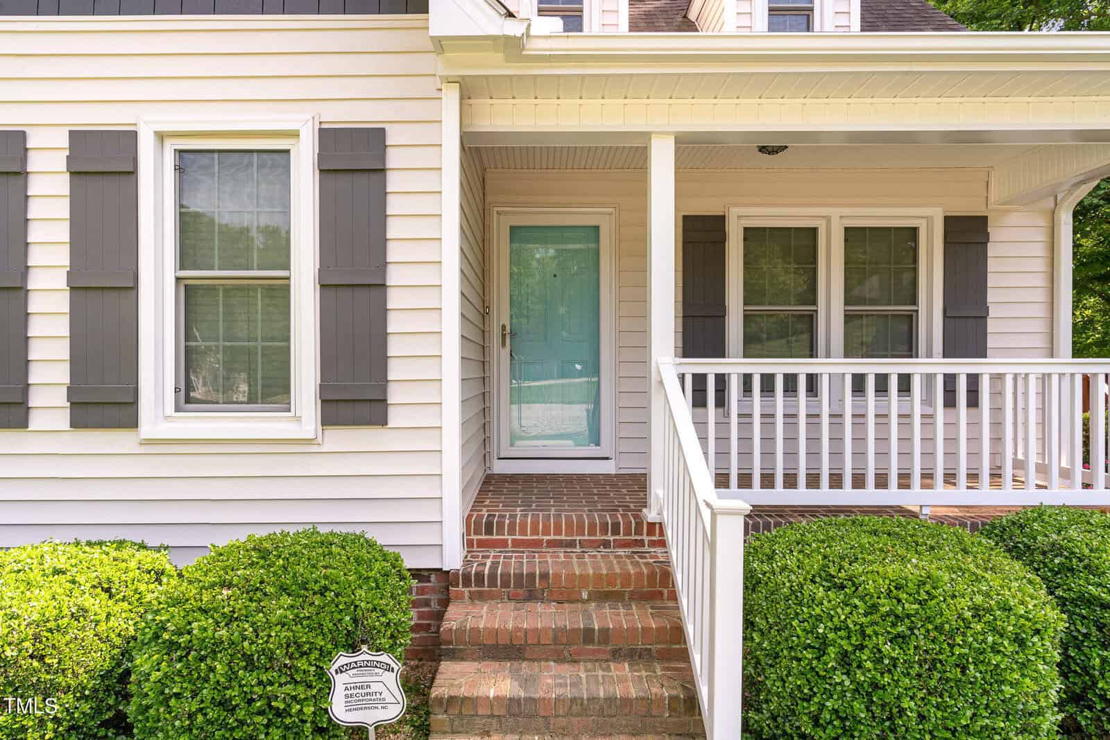A front porch with brick steps, white railing, and a light blue door. The house exterior is light-colored with gray shutters and multiple windows. Green shrubs line the walkway leading to the entrance.