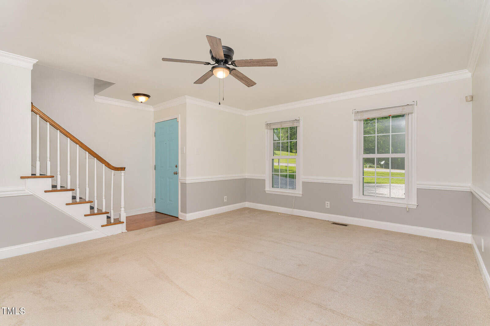 A bright living room with beige carpet, light gray and white walls, two windows, a ceiling fan with lights, and a staircase with wooden steps and white spindles. A blue front door is visible near the stairs.