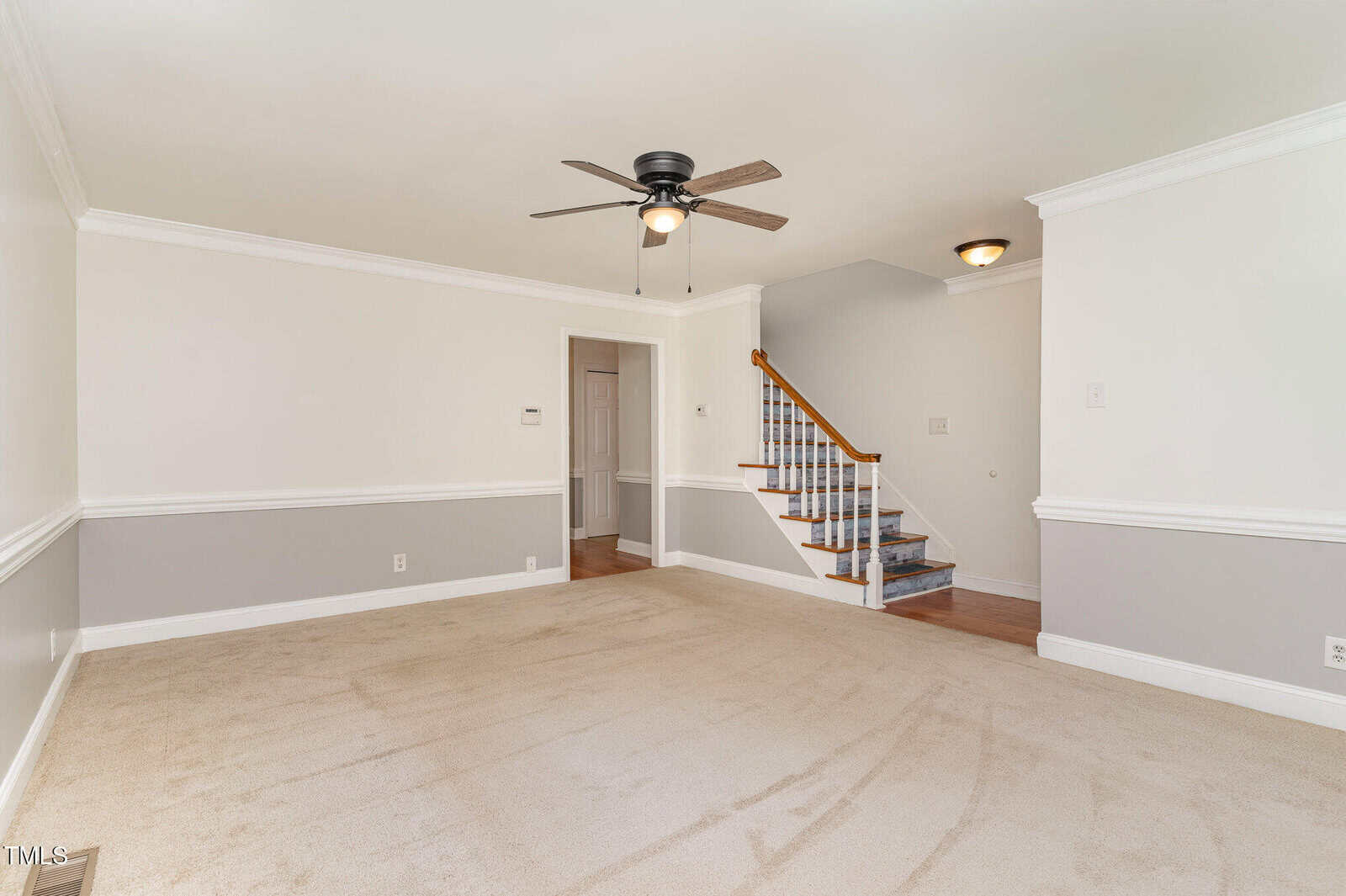 A spacious, empty living room features light beige carpet, two-tone white and gray walls with chair rail molding, a ceiling fan with light, and a staircase with a wooden handrail leading upward.