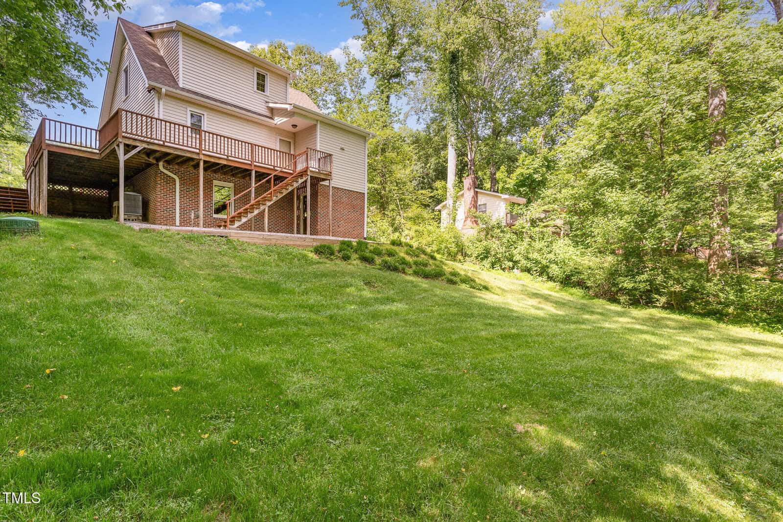 A two-story house with brick and siding exterior sits on a grassy, sloped yard surrounded by trees. A staircase leads to a wooden deck on the side of the house. Another house is visible in the background.