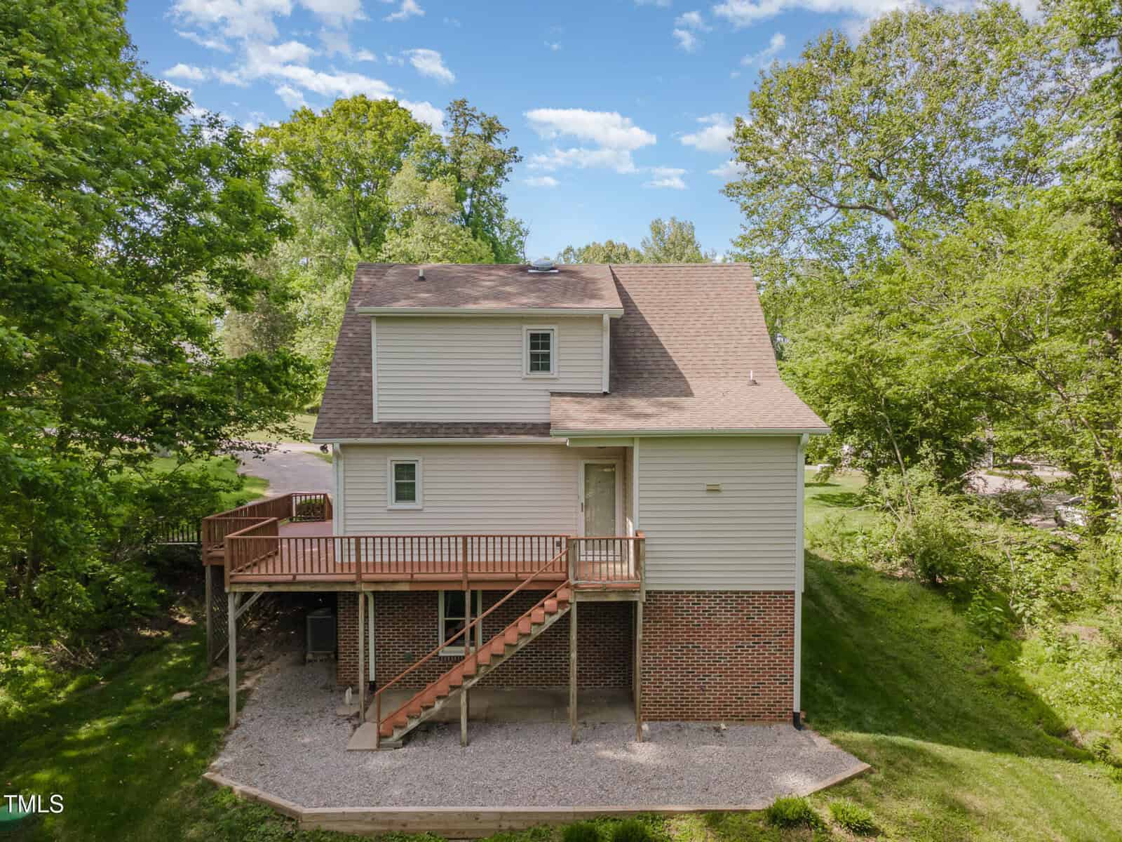 A two-story house with beige siding and red brick foundation, featuring a wooden deck and staircase, sits on a sloped grassy yard surrounded by trees under a partly cloudy sky.