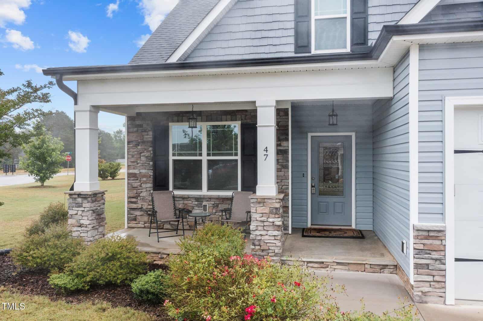Covered front porch of a gray house with stone accents, a small table and two chairs, a blue door with glass panel, and the number 47 displayed by the window. The porch overlooks a green lawn and landscaped garden.