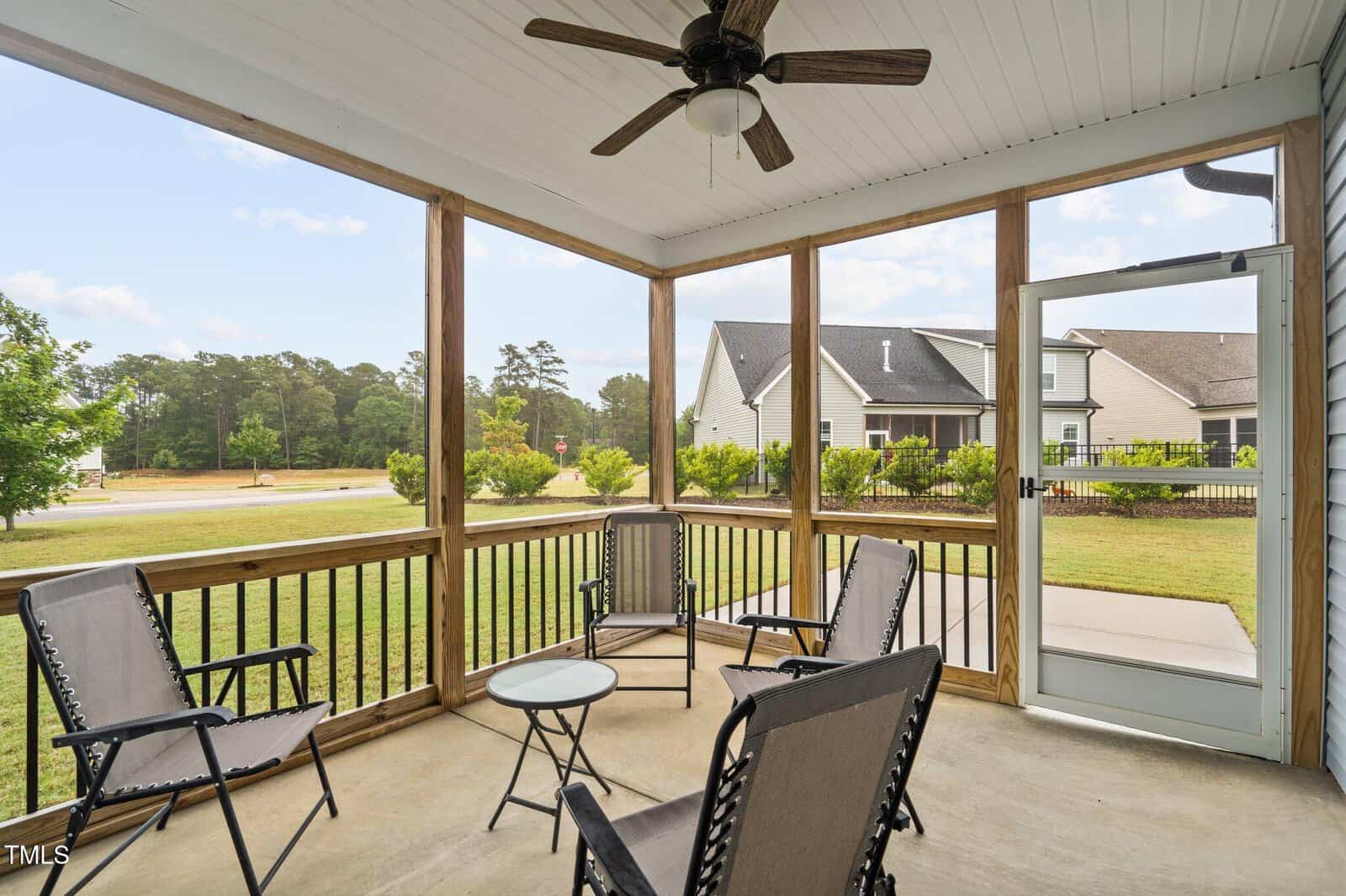 A screened-in porch with four patio chairs around a small round table, ceiling fan above, and views of a green lawn, nearby houses, and trees in the background.
