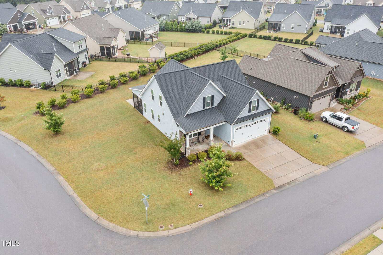 A two-story light gray house with a dark roof sits on a corner lot with a driveway and attached garage. Neighboring houses and lawns are visible, all separated by fences. Streets border the property on two sides.