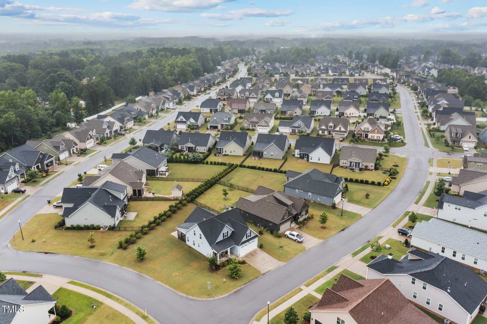 Aerial view of a suburban neighborhood with rows of single-family homes, well-maintained lawns, and curved streets. Houses have driveways and some cars are parked. Trees and forested area are visible in the background.