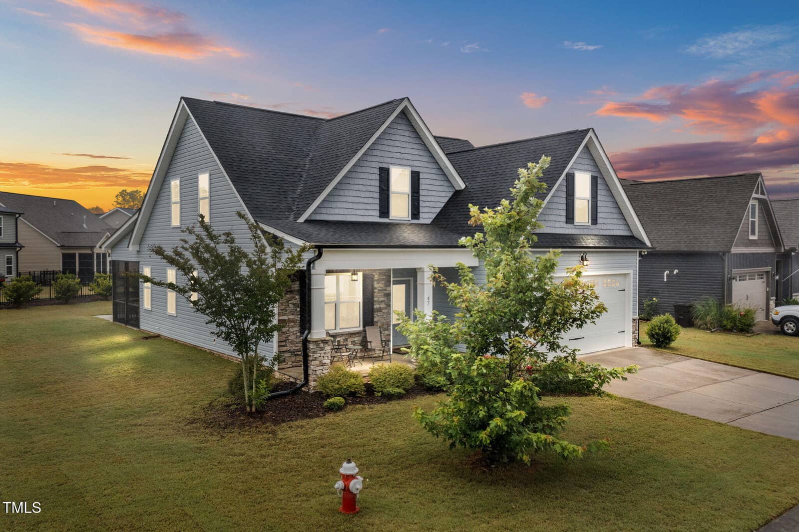 A modern two-story gray house with a front porch, well-kept lawn, and driveway at sunset. A small tree and red fire hydrant are in the front yard, with neighboring homes visible in the background.