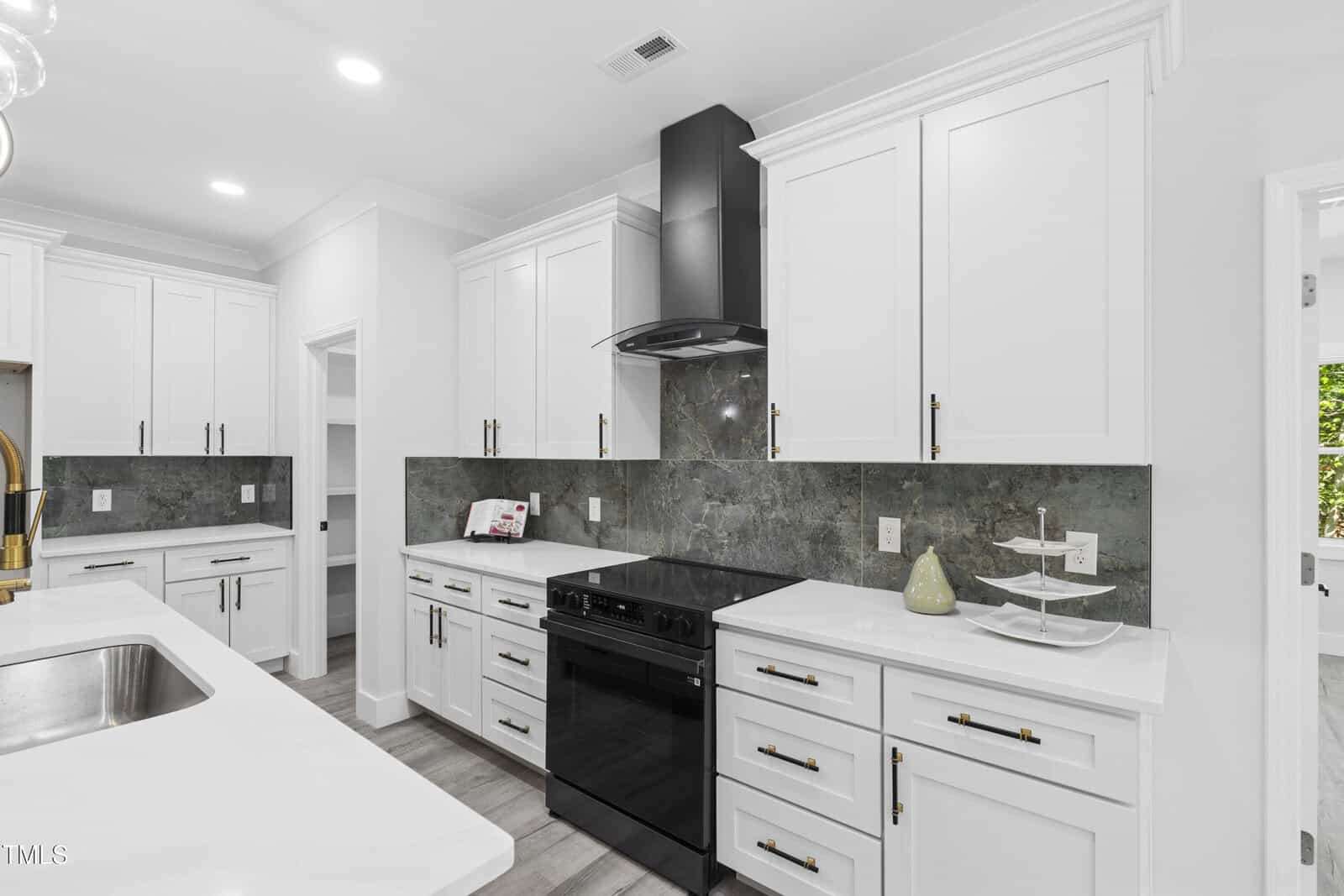 Modern kitchen with white cabinets, black stove and range hood, gray stone backsplash, white countertops, and a double sink. A tiered tray and decorative vase sit on the counter. Bright lighting and clean design.