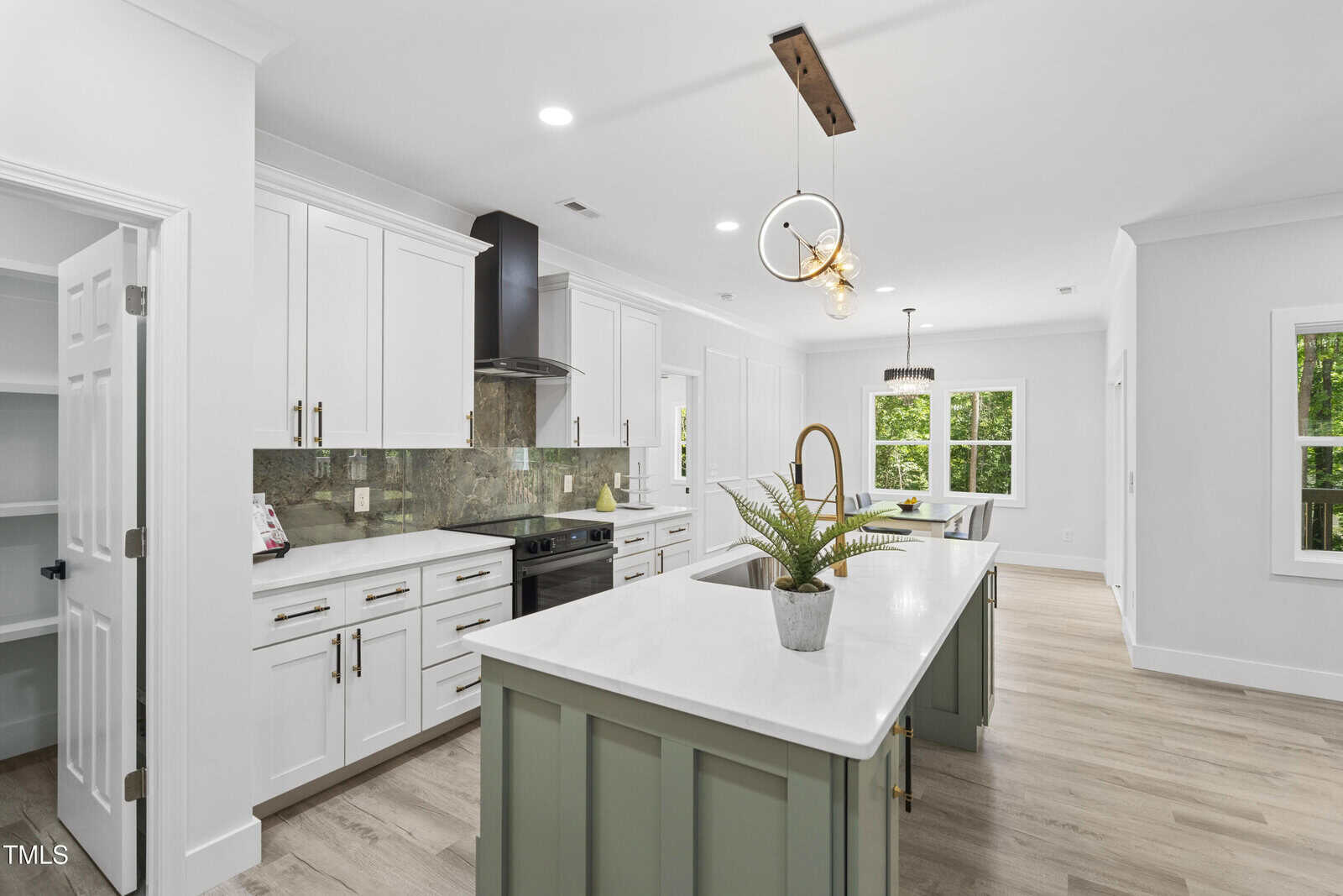 Modern kitchen with white cabinets, light green island, white countertops, stainless steel appliances, wood floors, a double pendant light, and a pantry with an open door. Large windows bring in natural light.