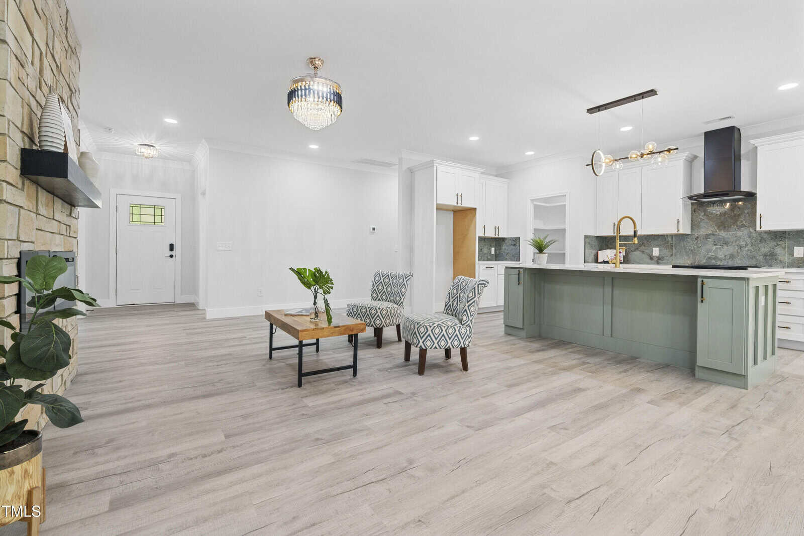 Open-concept living area with light wood floors, sage green kitchen island, white cabinets, stone accent wall, modern chandelier, two patterned chairs, coffee table, and potted plants; entryway visible in background.