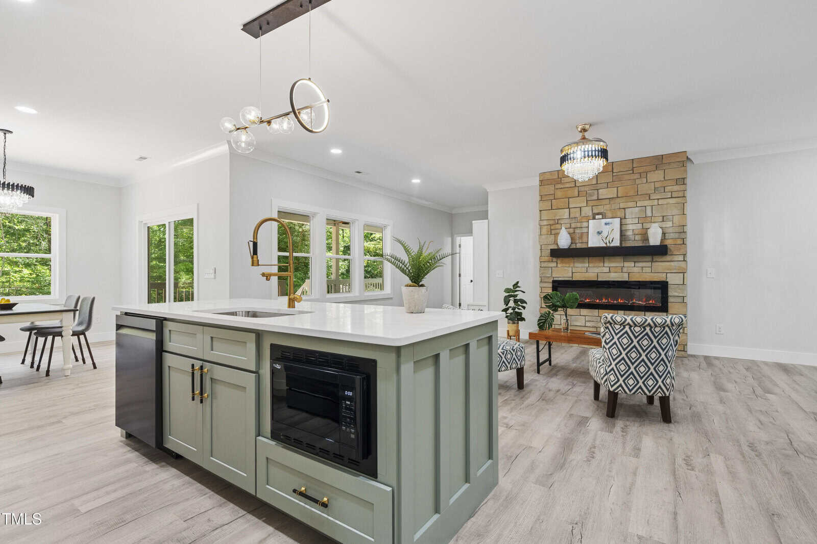 Modern open-concept kitchen and living area with a large island, gold faucet, and built-in microwave. Bright dining space, cozy seating, stone fireplace, and large windows with views of greenery.
