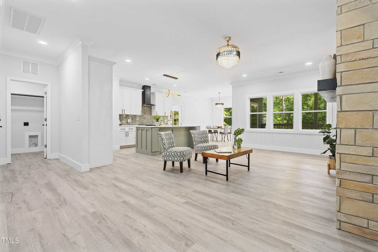 Open-concept living area with light wood flooring, modern kitchen with green island, white cabinets, dining table, large windows with green views, and a chandelier above two chairs and a coffee table.