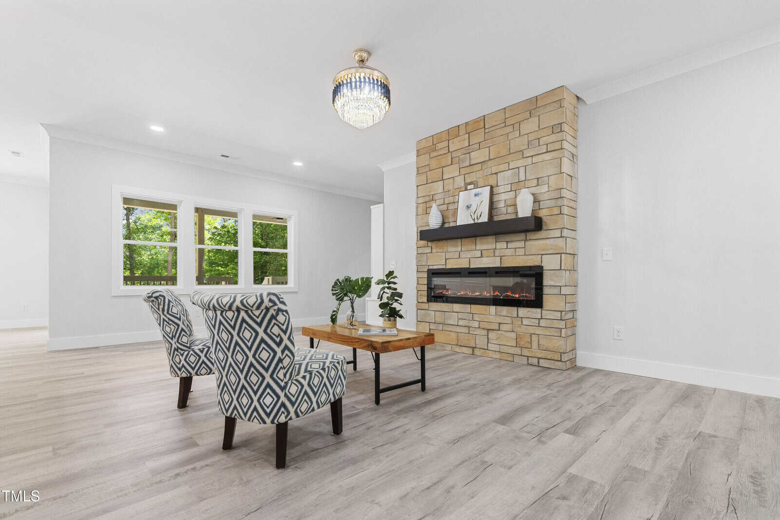 Modern living room with light wood floors, two patterned armchairs, a wooden coffee table with plants, stone fireplace with electric insert and two decorative items, large window, and a crystal chandelier.
