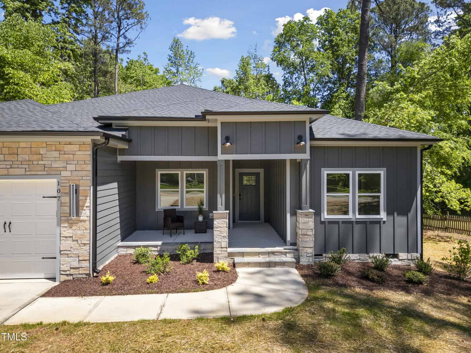 A modern single-story house with gray siding, stone accents, large windows, and a covered front porch. The home is surrounded by trees and has a landscaped front yard with a concrete walkway.
