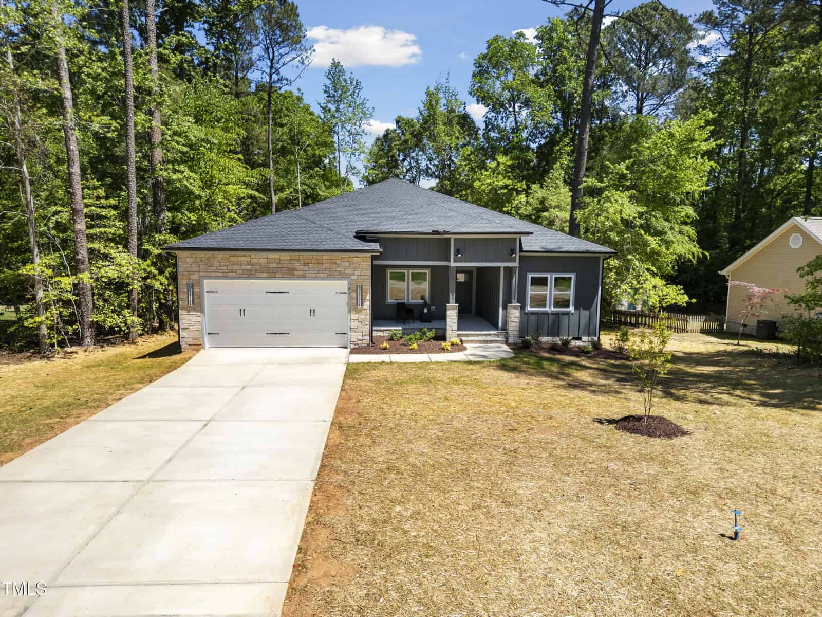 A modern single-story gray house with a two-car garage, front porch, and concrete driveway, surrounded by tall trees and a spacious yard with minimal landscaping under a blue sky.