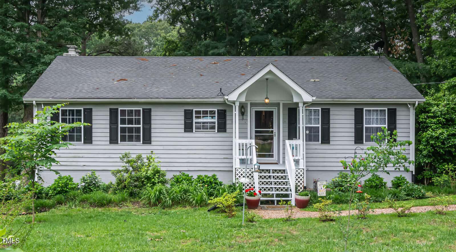 A small gray house with white trim, black shutters, and a covered front porch with steps and railings. The yard features green grass, shrubs, plants, and two potted flowers by the porch. Trees surround the house.