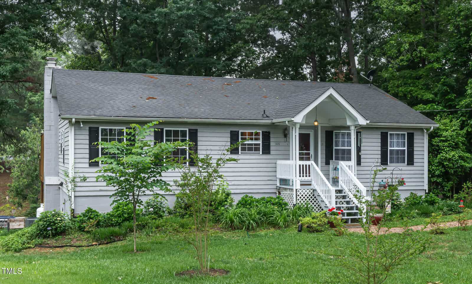A small gray house with white trim and a covered front porch, surrounded by a lush green lawn and trees, with garden plants and a stone path leading to the entrance.
