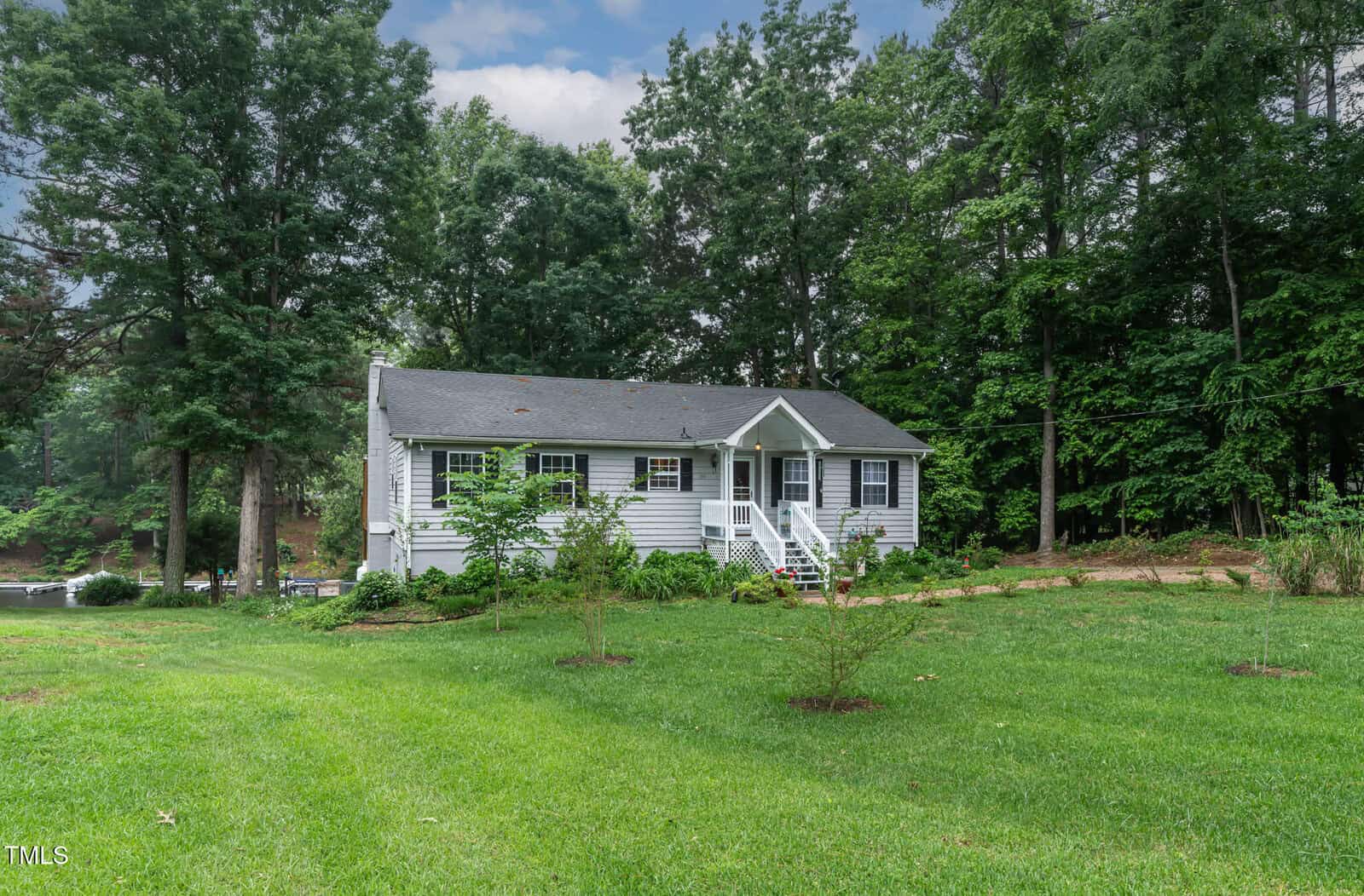 A small, light gray house with white trim sits in front of a large green lawn, surrounded by tall trees and shrubs under a partly cloudy sky. Steps lead up to a covered front porch.