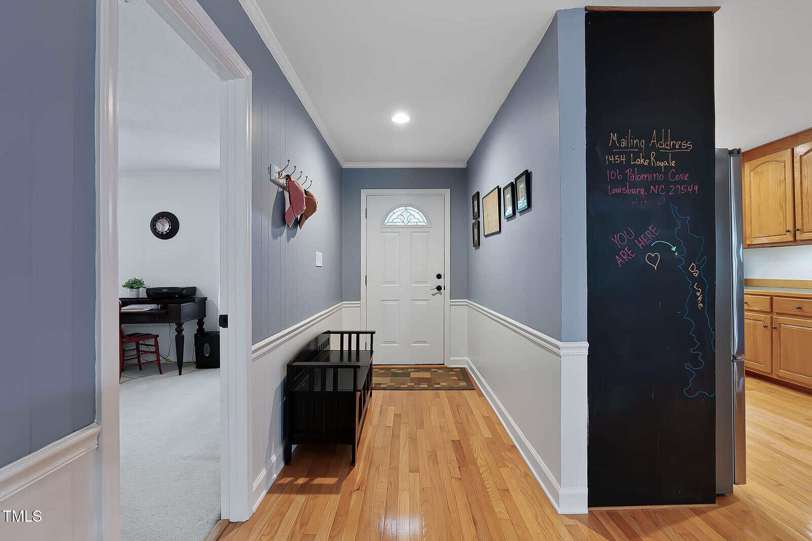 A hallway with light hardwood floors, blue-gray walls with white trim, a white front door, wall hooks with bags, a black bench, framed photos, and a chalkboard wall with notes next to a kitchen area with wooden cabinets.