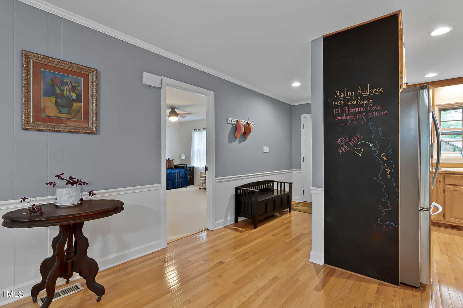 A hallway with gray and white walls, light wood floors, a small table with a plant, a wall bench with hooks above, and a black chalkboard wall with writing next to an entry to the kitchen.