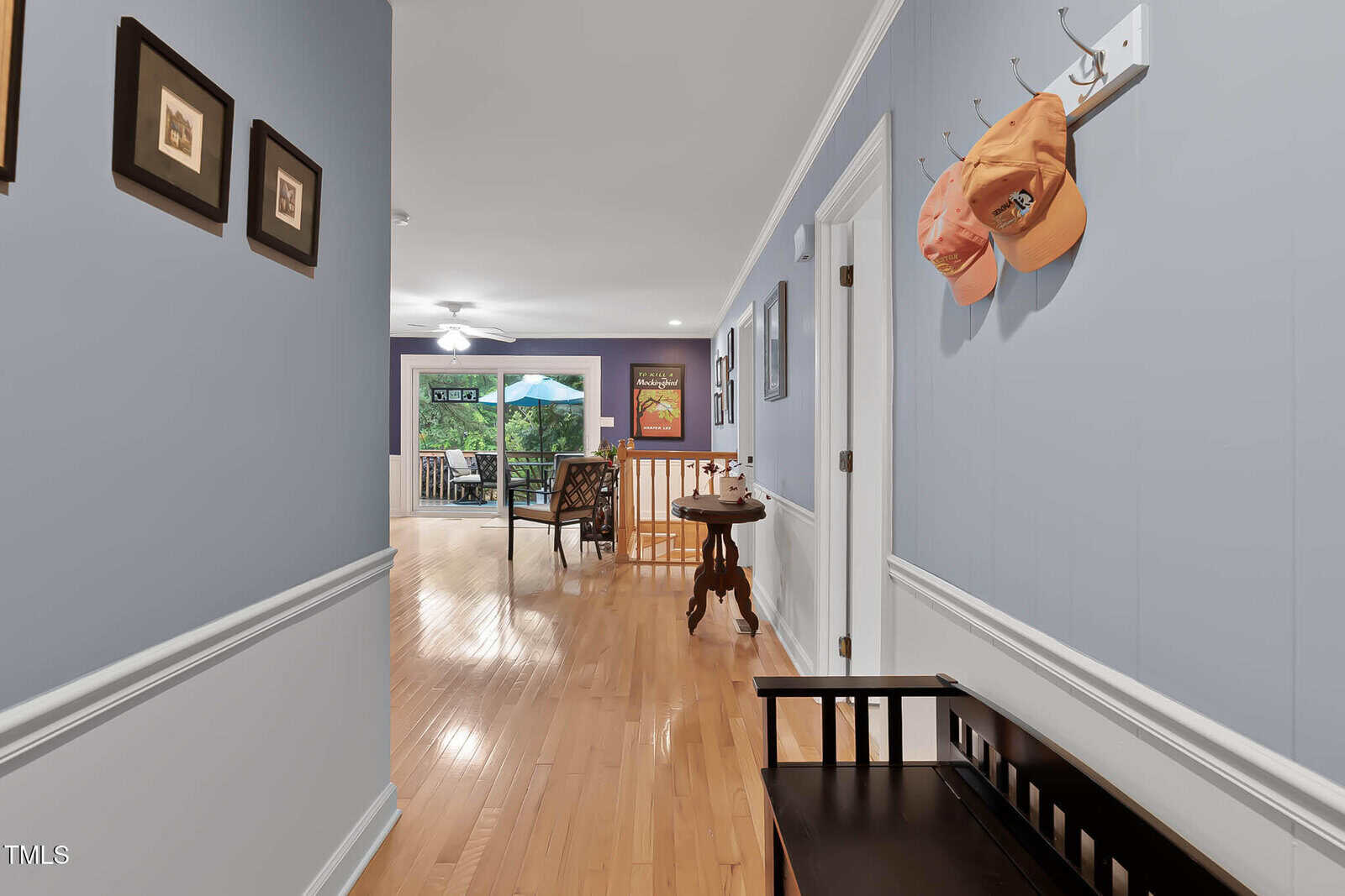 A hallway with light purple walls and wood flooring leads to a living area with a ceiling fan. Two orange hats hang on hooks above a black bench. Framed art is on the walls. A patio with an umbrella is visible outside.