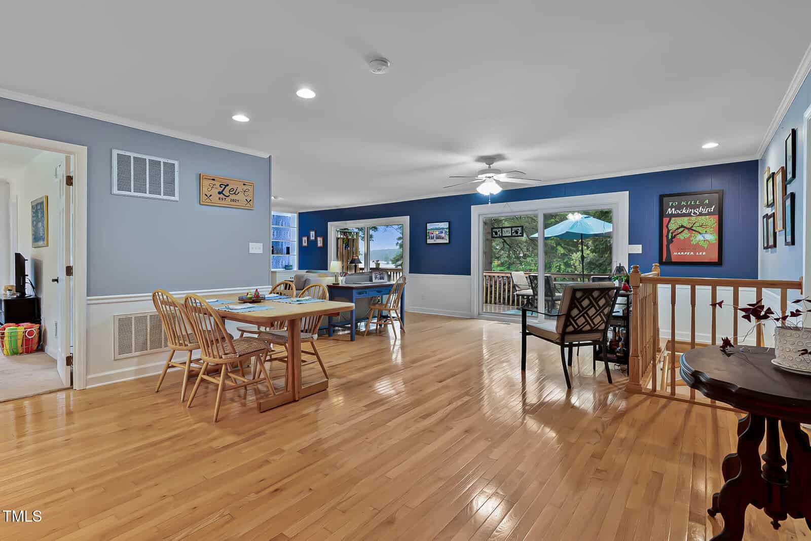 Open dining and living area with wood flooring, blue walls, a dining table with chairs, a ceiling fan, wall art, and sliding glass doors leading to a deck with a view of greenery outside. Kitchen is visible in the background.