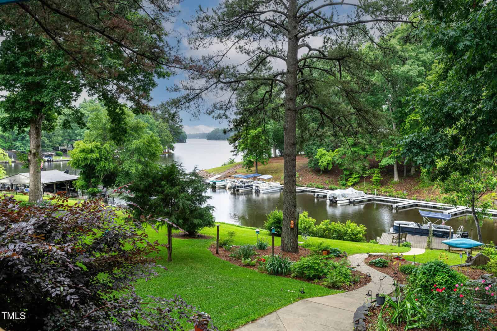 A lush green backyard with a winding stone path leads to a small dock on a calm lake, surrounded by trees and several boats moored at the shore under an overcast sky.