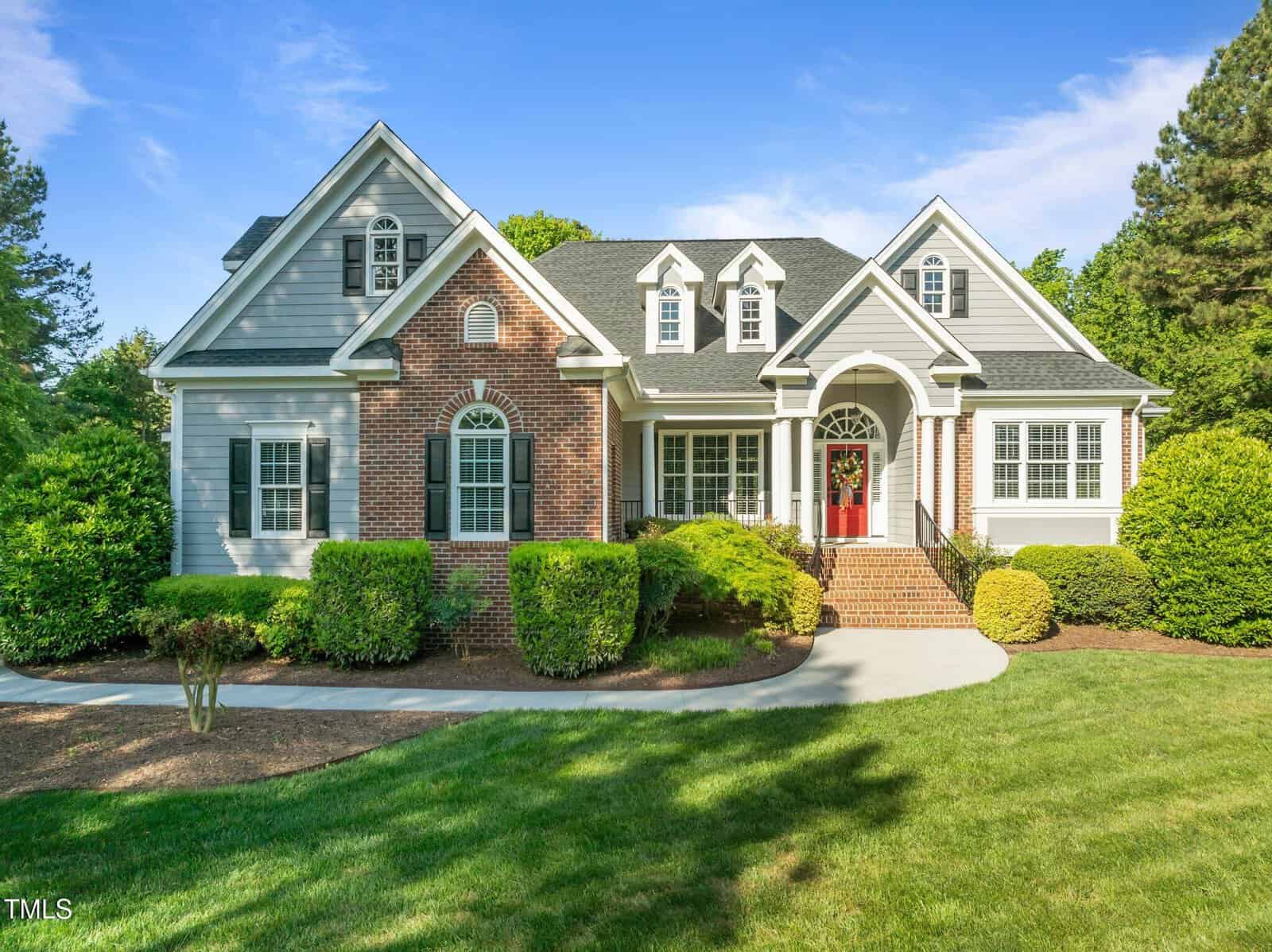A two-story suburban house at 503 Belmont Circle in Wake Forest with gray siding and red brick accents, white trim, multiple front-facing gables, a red front door, and a well-kept lawn with shrubs and trees under a clear blue sky.