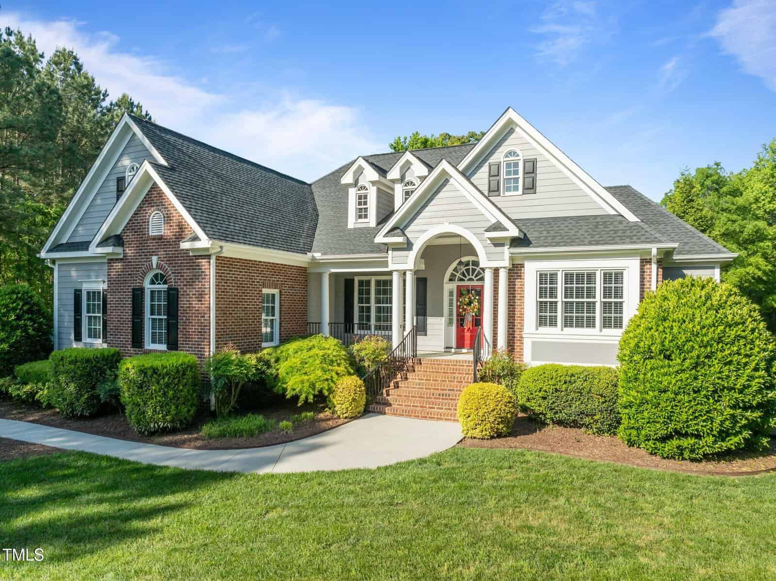 Single-story suburban house with a brick and white siding exterior, arched entryway, red front door, multiple gables, manicured lawn, landscaped bushes, and a curved walkway under a clear blue sky.