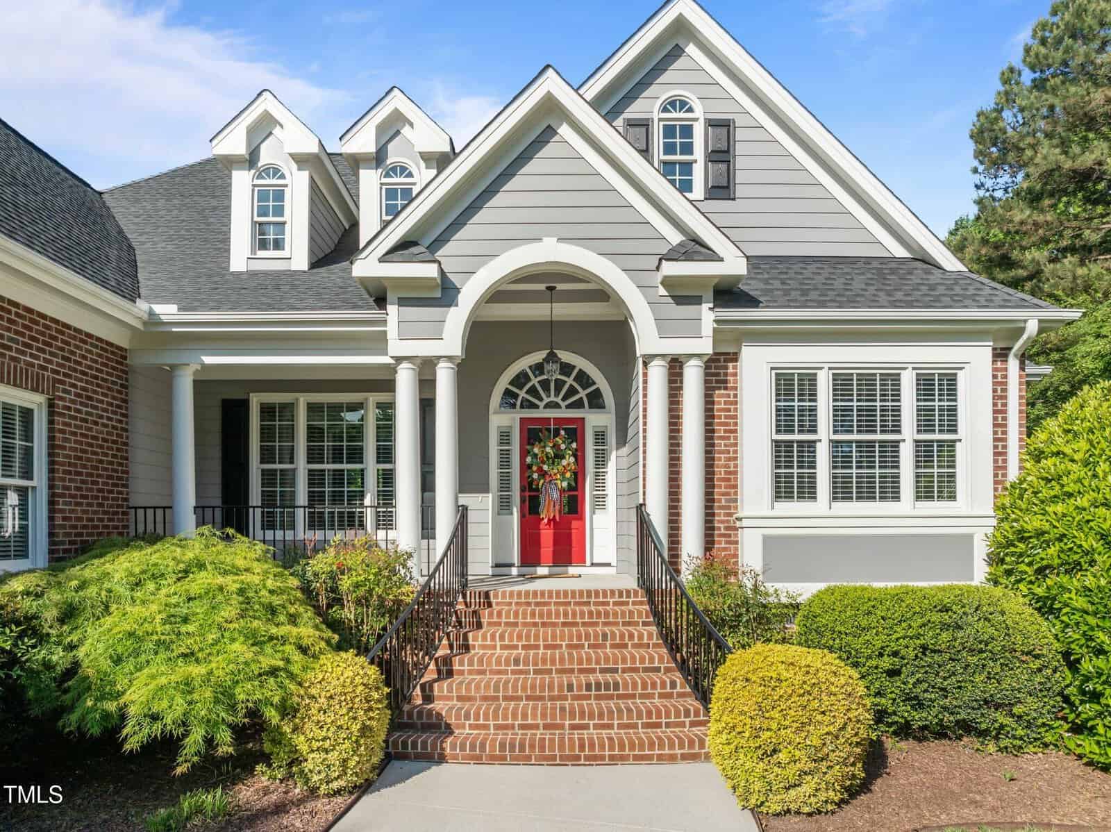 A two-story house with gray siding, brick accents, and white trim. It has a red front door, arched entryway, double staircase, dormer windows, and landscaped bushes in front.