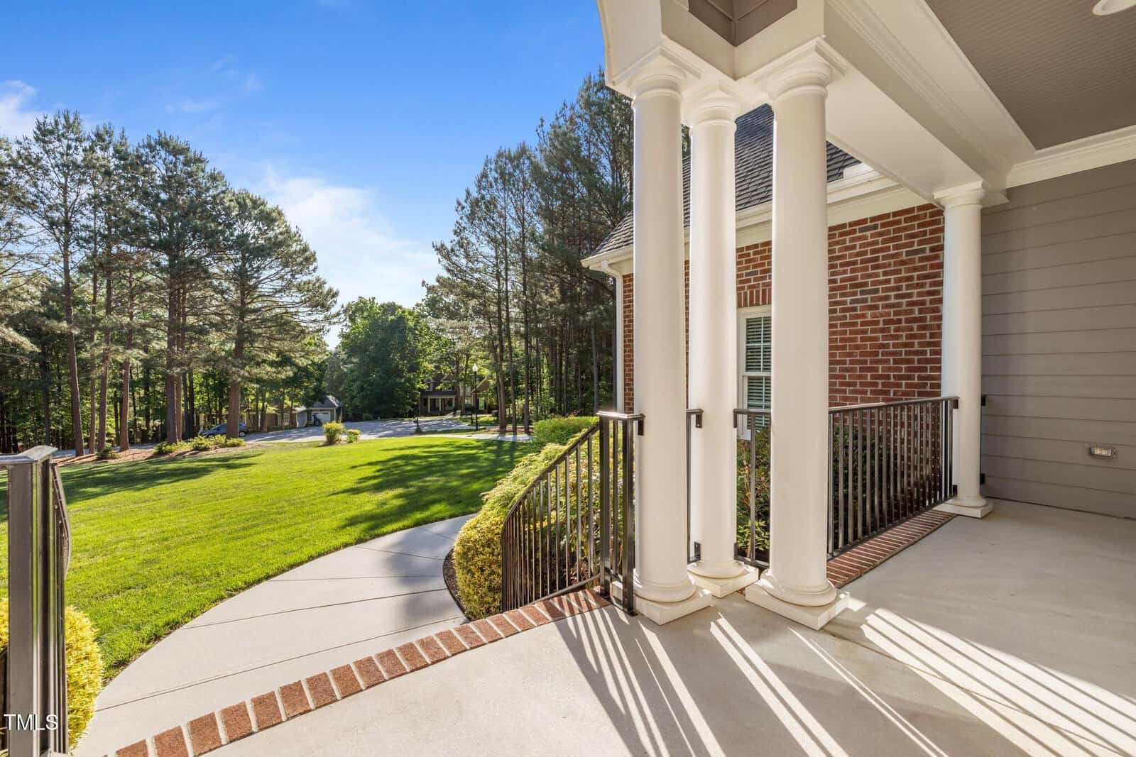 A view from a covered front porch with white columns and black railings, overlooking a well-maintained lawn, curved walkway, and a wooded area with tall trees under a clear blue sky.