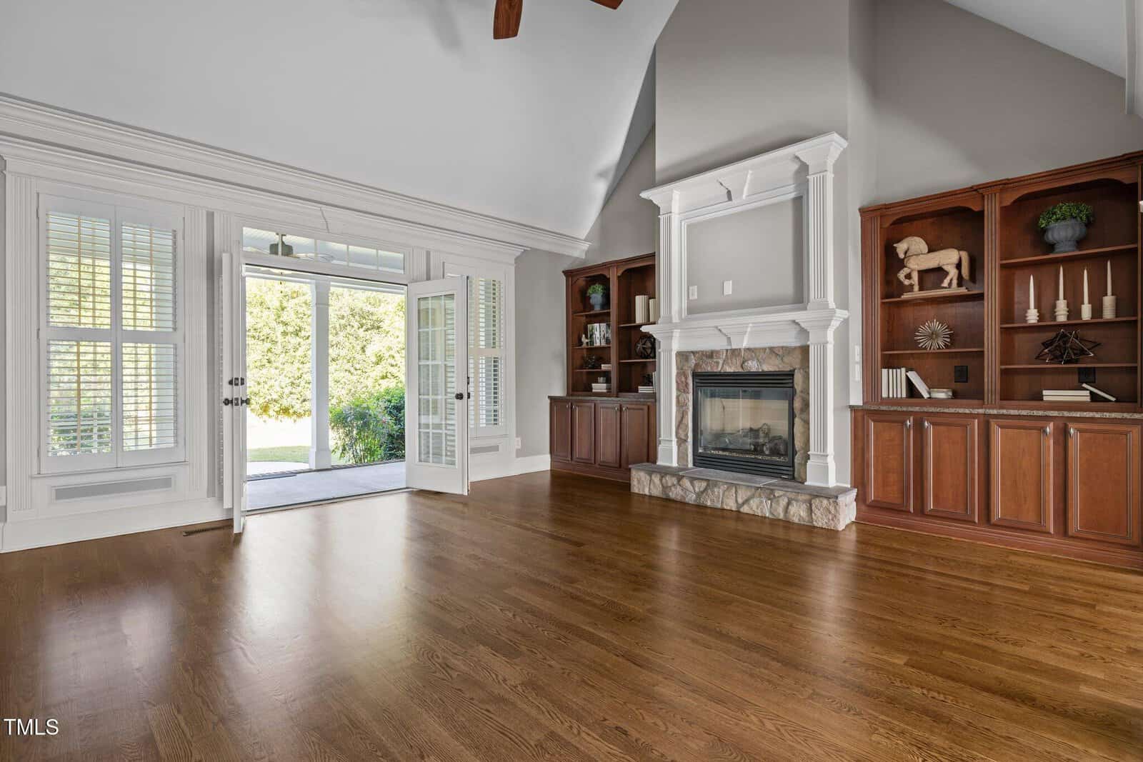 Spacious living room with hardwood floors, built-in wooden shelves and cabinets, a fireplace with a white mantle, and double glass doors leading to a green outdoor area. White plantation shutters cover the windows.