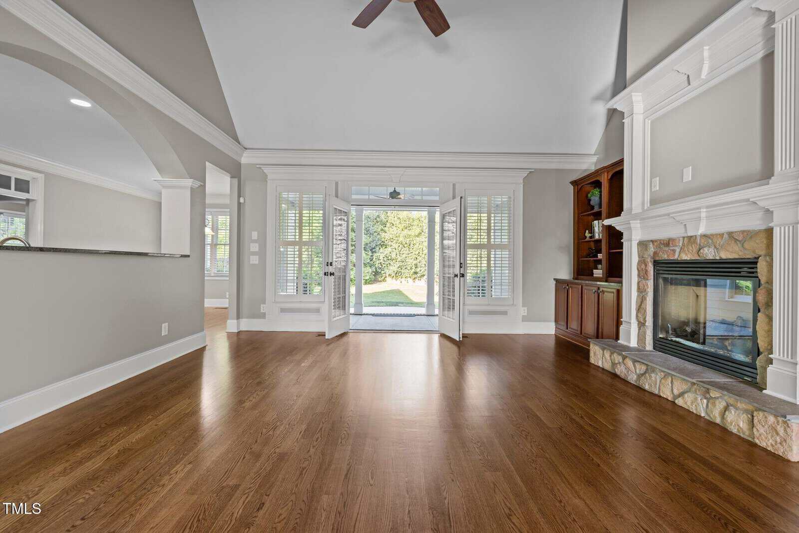 A spacious living room with hardwood floors, a stone fireplace with a white mantel, built-in shelves, and double glass doors opening to a bright sunroom with views of greenery outside.
