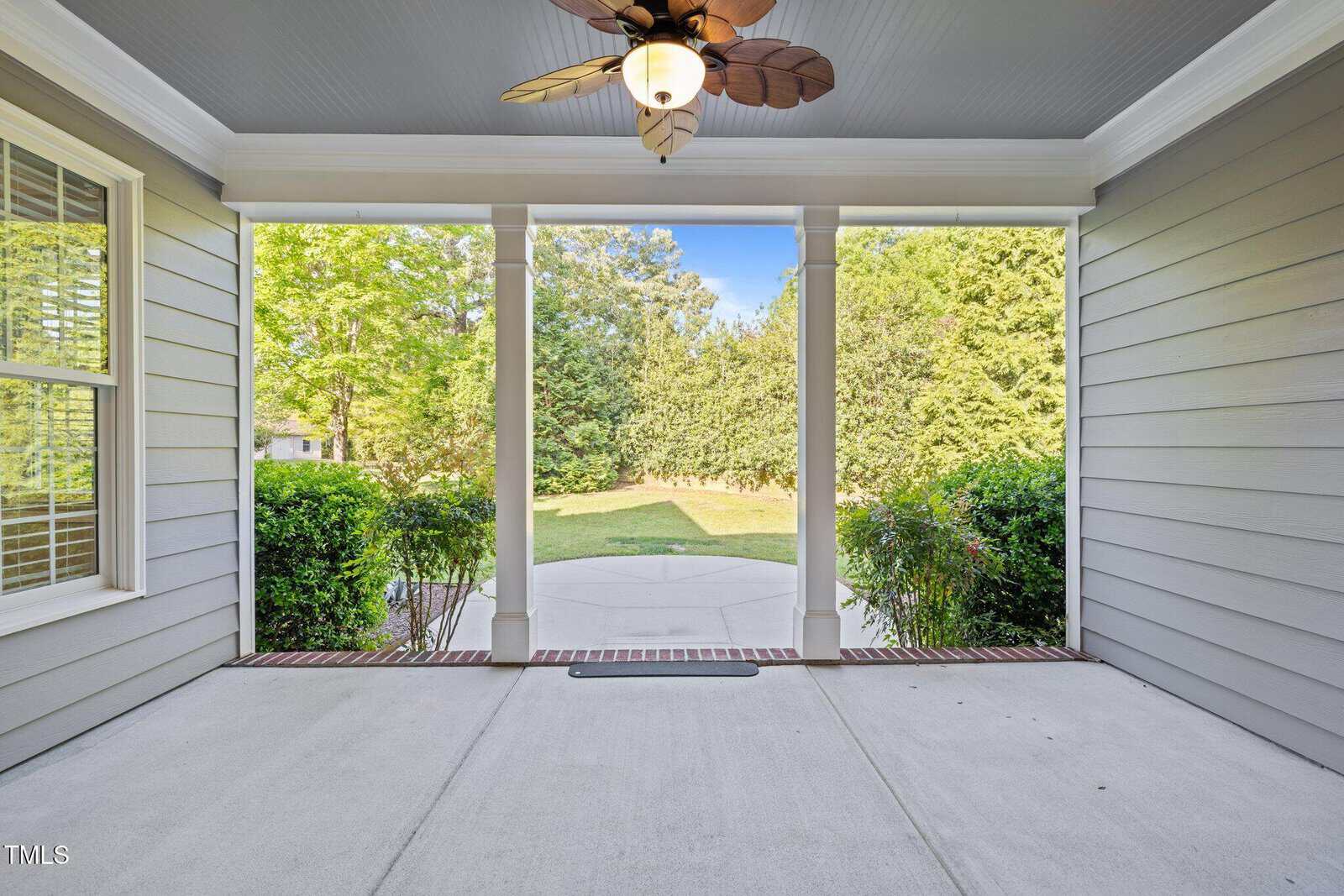 View from a covered porch with a ceiling fan and columns, looking out onto a concrete patio, green shrubs, and a grassy yard bordered by trees under a clear sky.
