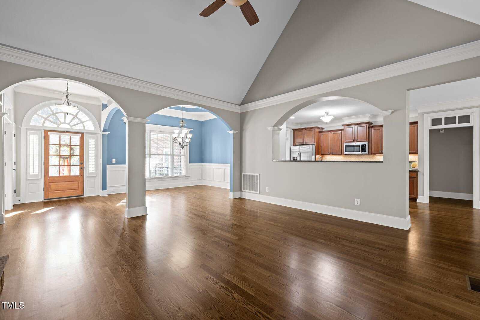 Open-plan living area with hardwood floors, vaulted ceiling, and neutral walls. Large entryway features wooden double doors and an arched transom window. Visible kitchen, dining area, and arched openings.