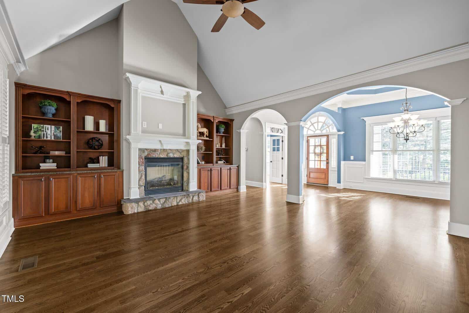 Spacious living area with hardwood floors, built-in wooden shelves, a fireplace with a stone base, vaulted ceiling with fan, arched doorway, and large windows letting in natural light.